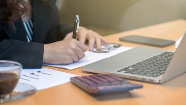 mulher fazendo contas e escrevendo em papéis em frente ao computador