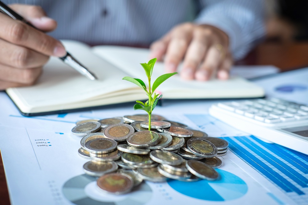 pilha de moedas em cima de papéis com resultados em gráficos de investimentos e ao fundo homem escrevendo