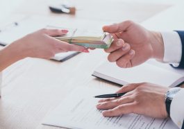duas mãos, uma dando dinheiro à outra, junto a papéis de contrato