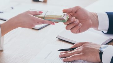 duas mãos, uma dando dinheiro à outra, junto a papéis de contrato