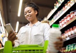moça jovem mexendo no celular enquanto segura um carrinho de compras no supermercado