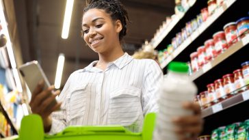 moça jovem mexendo no celular enquanto segura um carrinho de compras no supermercado