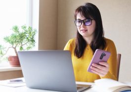 mulher de óculos mexendo no computador e com o celular em mãos