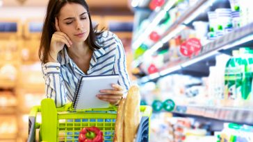 mulher olhando desconfiada para a lista de compras apoiada em um carrinho de compras no supermercado