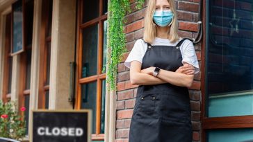 mulher de máscara em frente à um restaurante com a placa de fechado, indicando a crise de desemprego no Brasil