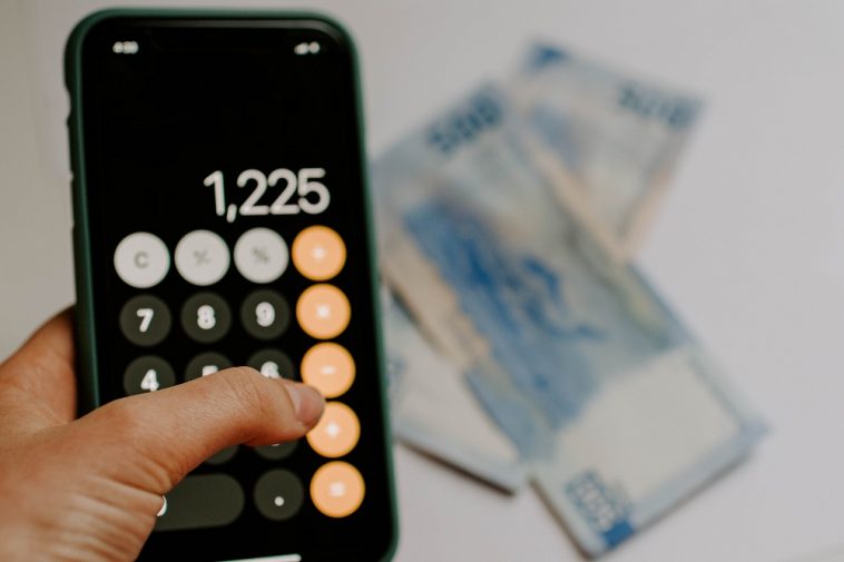 mão de mulher calculando conta na calculadora do celular e notas de dinheiro ao fundo