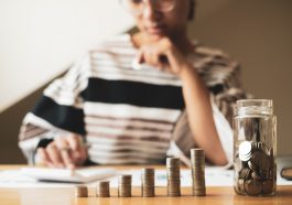 mulher ao fundo desfocado calculando na calculadora e a frente pilhas crescentes de moeda simbolizando guardar e investir dinheiro