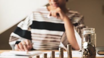 mulher ao fundo desfocado calculando na calculadora e a frente pilhas crescentes de moeda simbolizando guardar e investir dinheiro