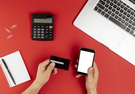 mão feminina segurando cartão de crédito e mexendo no celular, ao lado um bloco de anotações e calculadora