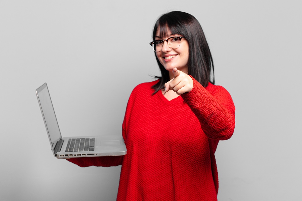 mulher em pé, segurando um notebook aberto e apontando o dedo para você