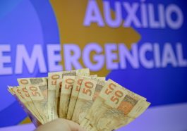 mão de pessoa com notas de cinquenta reais ao fundo de uma imagem escrita auxílio emergencial, simbolizando opagamento da quinta parcela do auxílio emergencial