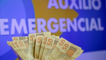 mão de pessoa com notas de cinquenta reais ao fundo de uma imagem escrita auxílio emergencial, simbolizando opagamento da quinta parcela do auxílio emergencial