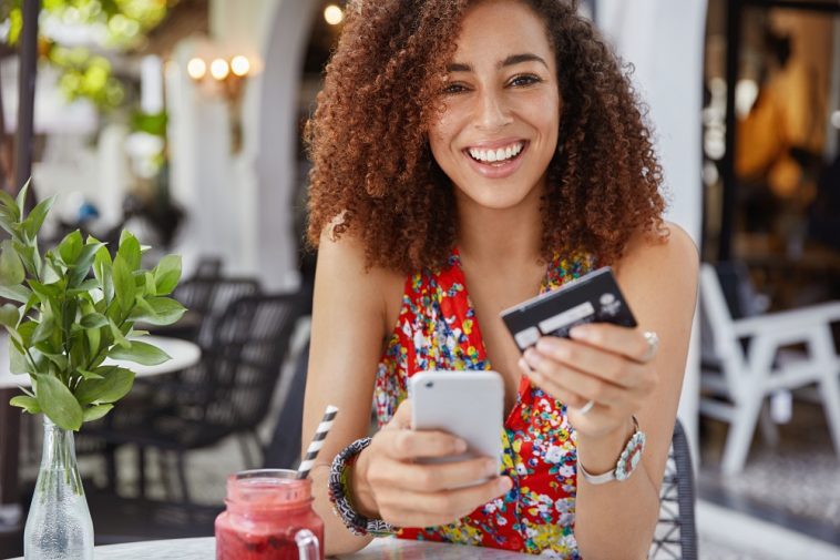 mulher jovem feliz e sorridente com penteado afro, usa um celular moderno e um cartão de crédito para fazer compras online