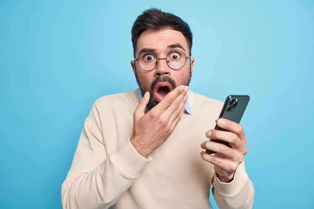 Homem europeu adulto barbudo estupefato mantém a boca aberta e olha fixamente aterrorizado, segura o celular descobre notícias chocantes vestido com um macacão casual