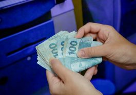 mão feminina contando notas de cem em frente à um caixa eletrônico, simulando o recebimento do 14° salário do INSS