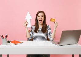 mulher sentada em uma mesa em escritório de casa com uma mão o cartão de crédito e na outra uma seta apontando para cima, com cara de felicidade, simbolizando a descoberta dos Melhores cartões de crédito com aumento de limite atrelado a investimento