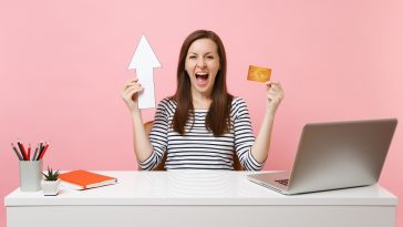 mulher sentada em uma mesa em escritório de casa com uma mão o cartão de crédito e na outra uma seta apontando para cima, com cara de felicidade, simbolizando a descoberta dos Melhores cartões de crédito com aumento de limite atrelado a investimento