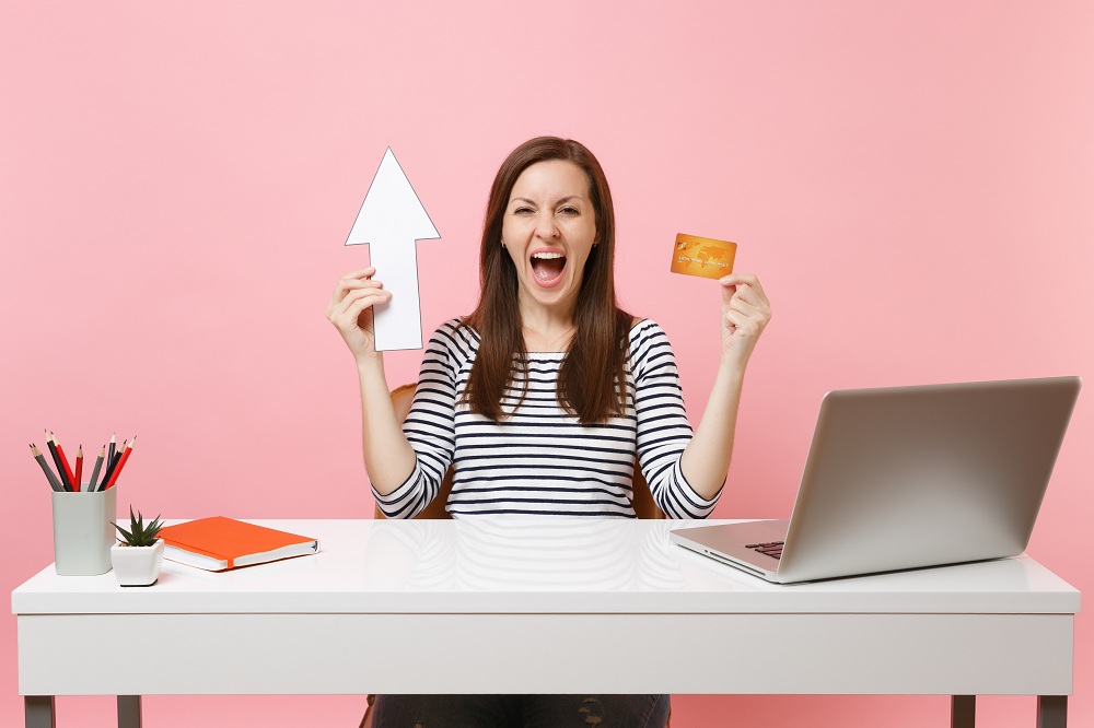 mulher sentada em uma mesa em escritório de casa com uma mão o cartão de crédito e na outra uma seta apontando para cima, com cara de felicidade, simbolizando a descoberta dos Melhores cartões de crédito com aumento de limite atrelado a investimento