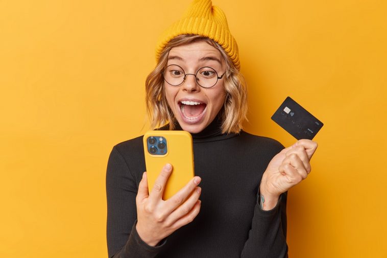Mulher feliz com semblante de surpresa olhando para o celular que está com a tela do aplicativo com conta digital gratuita e na outra mão cartão de crédito