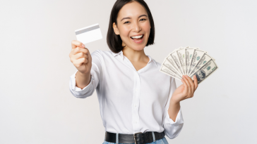 mulher coreana feliz e sorridente segurando cartão em uma mão e na outra notas de dólar, simbolizando como ganhar dinheiro com o cartão de crédito