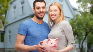 casal jovem feliz segurança cofrinho de porquinho ao fundo de uma casa, simbolizando Como juntar dinheiro em casal