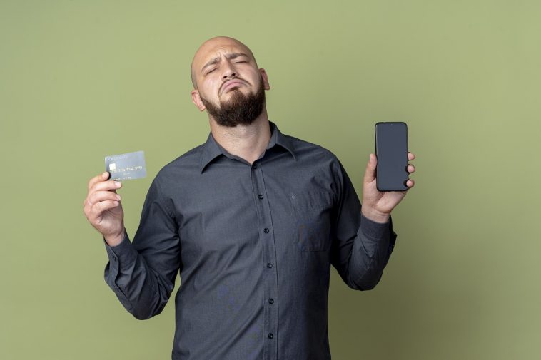Homem jovem e triste, careca, segurando um telefone celular e um cartão de crédito com os olhos fechados, simbolizando um cartão de crédito descontinuado, cancelado, fim