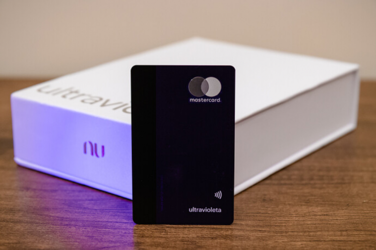 imagem do cartão de crédito Nubank Ultravioleta Mastercard Black a frente da caixa de boas vindas, com uma luz roxa UV ao fundo