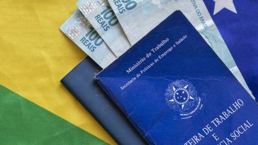 carteira de trabalho do Brasil e notas de cem reais dentro dela e ao fundo uma bandeira do Brasil, para simbolizar o novo valor do salário mínimo para 2023