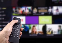Mão segurando o controle remoto de tv com smart tv ligada em plataforma de streaming, simbolizando o acesso a Netflix mais barata no plano básico com anúncios