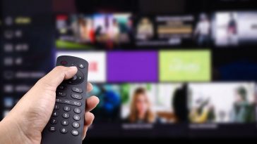 Mão segurando o controle remoto de tv com smart tv ligada em plataforma de streaming, simbolizando o acesso a Netflix mais barata no plano básico com anúncios