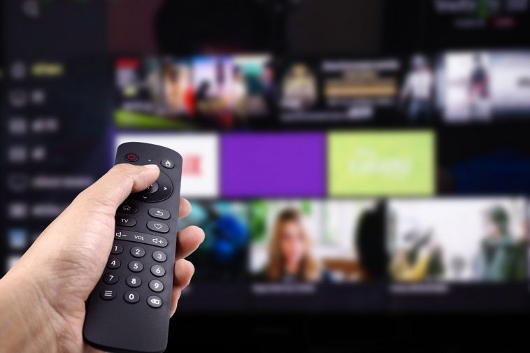 Mão segurando o controle remoto de tv com smart tv ligada em plataforma de streaming, simbolizando o acesso a Netflix mais barata no plano básico com anúncios